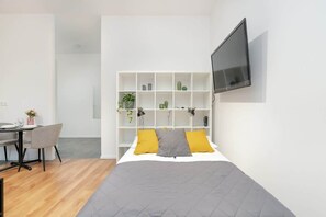 A bright bedroom area with a double bed, grey and yellow bedding, and minimalist decor, including a wall-mounted TV.
