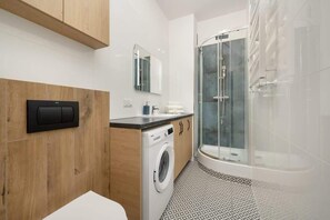 A bathroom corner featuring a washing machine, wooden storage, and a glass-enclosed shower cabin.
