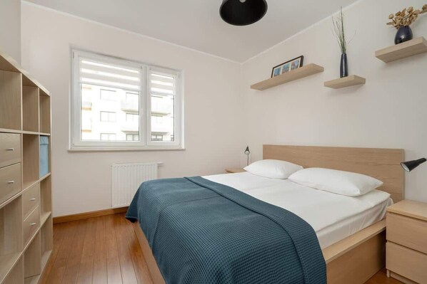 A cozy bedroom with a double bed, white and blue bedding, wooden accents, and soft lighting, creating a comfortable atmosphere.
