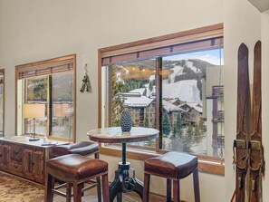 A cozy indoor setting of a room with high ceilings, featuring a small round table and two stools near a large window showcasing a view of snow-covered buildings and mountains outside. Skis are propped against the wall.