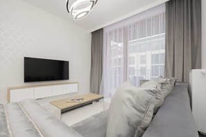 A bright entertainment space with a mounted TV, a sleek white media unit, and floor-to-ceiling windows letting in natural light.

