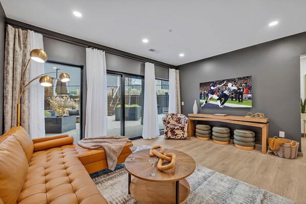 Modern living room decorated in calming tones, featuring comfortable sectional and flat-screen smart TV, and is connected to the kitchen.