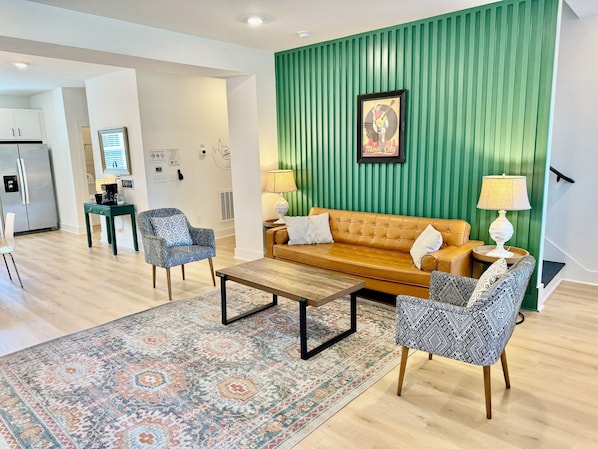 LIVING ROOM – This vibrant living room is styled with a mix of modern and vintage elements, creating a cozy yet contemporary atmosphere. The bold green accent wall features iconic local artwork, adding a touch of Nashville's character to the space. A mustard-yellow leather sofa and patterned chairs invite you to relax in style, while the nearby kitchen and dining area provide a...