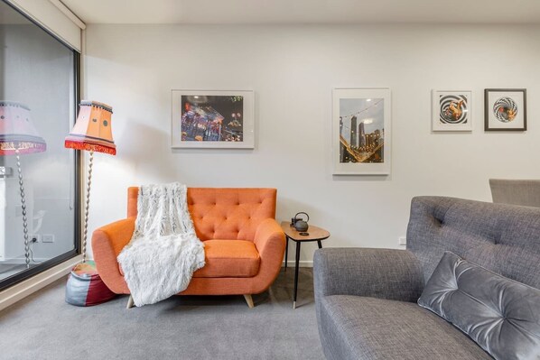 A living area is furnished with an eclectic orange couch, quirky artwork and an additional grey armchair