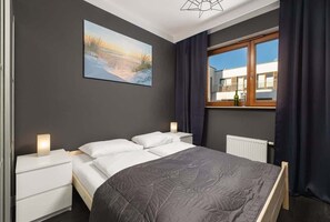 Bedroom with a wardrobeA spacious bedroom with a double bed next to which are white bedside tables with lamps. Above the bed hangs a painting with a seaside landscape. Long curtains hang in the window. The walls are grey in colour. 