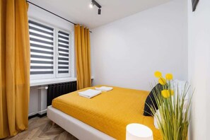 A cozy bedroom featuring a comfortable bed with mustard yellow bedding, a black bedside table, and large windows with blinds.
