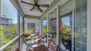 A small screened-in balcony with a ceiling fan, three wicker chairs with cushions, a wooden table, and surrounding lush greenery outside. Large glass doors provide access to the interior.