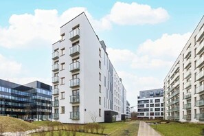 A view of the apartment complex from the outside, showcasing the modern architecture with clean lines and light-colored facades. Surrounding greenery and well-maintained paths add to the welcoming atmosphere.