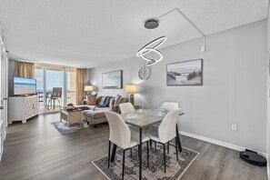 This beautifully designed open-plan living and dining area features a seamless blend of comfort and style. A modern dining table with white chairs sits under a striking spiral light fixture, setting the tone for sophisticated dining.