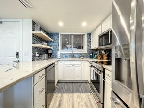 A bright, open kitchen with white cabinetry, marble countertops, and modern appliances, accented by blue tile backsplash and open shelving for a functional yet elegant space.
