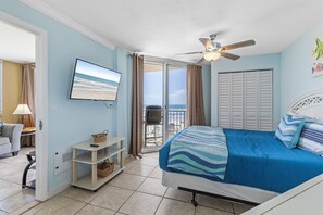 This bright bedroom offers a comfortable bed, wall-mounted TV, and direct access to a private balcony with stunning ocean views. Relax in coastal comfort with soft blue tones and a ceiling fan for added breeze.