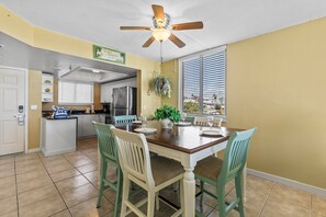 This charming dining space offers a comfortable setting with ample sunlight through large windows. The hanging plant and light fixtures add a relaxed, tropical vibe, while the open layout leads to a fully-equipped modern kitchen.