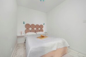 A cozy bedroom featuring a pastel pink upholstered headboard, a white bed, and minimalist decor.
