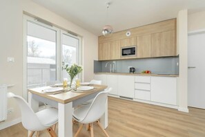 A modern open-plan kitchen and dining space equipped with light wood cabinets, a white dining table, and broad windows offering ample daylight.

