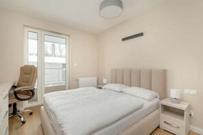 A cozy bedroom featuring a double bed with a neutral headboard, a bedside table, and a soft overhead light. A window provides a pleasant outdoor view.