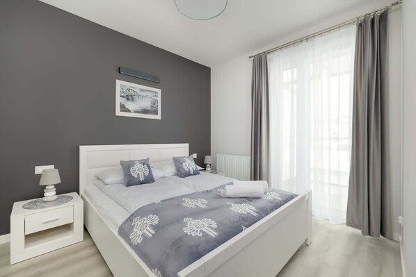 A stylish bedroom with a gray upholstered bed, white bedside tables, and floor-length curtains.
