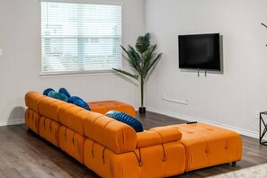 Bright living room featuring a bold orange sectional sofa with cozy pillows, perfect for lounging. A wall-mounted TV provides entertainment, making this space ideal for relaxing with family or friends after a long day.