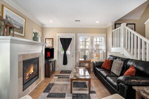 A cozy living room features a black leather sofa with orange and grey throw pillows, positioned next to a marble-topped coffee table. A fireplace with a white mantle. A staircase with white railings leads upstairs, adding to the room's charm.