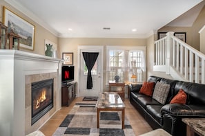 A cozy living room features a black leather sofa with orange and grey throw pillows, positioned next to a marble-topped coffee table. A fireplace with a white mantle. A staircase with white railings leads upstairs, adding to the room's charm.