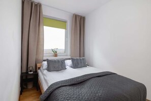 A cozy bedroom with a neatly made double bed, soft gray bedding, and a window with beige curtains, creating a peaceful sleeping space.

