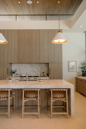 This modern, minimalist kitchen features warm, neutral tones with light wood cabinetry and a marble countertop. The spacious layout includes a central island with wicker stools, pendant lighting, and subtle decor for a clean, elegant look.