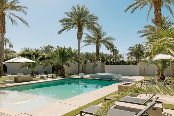 The backyard of this luxury estate features a large pool with a jacuzzi and waterfall, surrounded by a spacious patio. Enjoy dining, lounging, and a covered outdoor kitchen, all set in a tropical oasis with lush greenery and modern architecture.