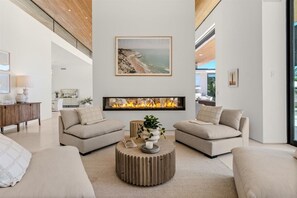 This modern living room features a high ceiling and a sleek fireplace as the focal point. Two beige sofas surround a wooden coffee table, while beach-themed art adds color. Minimalist decor and neutral tones create a serene, inviting space.