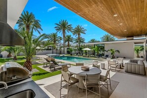 This luxurious desert estate features a large swimming pool surrounded by a lush lawn and tall palm trees. Relax on the patio with outdoor furniture, enjoy the shaded pergola, or cook on the built-in grill under a clear blue sky.