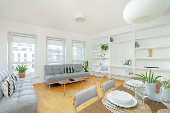 A bright and modern living room with a gray sofa, a round dining table set for meals, and large windows that allow plenty of natural light.

