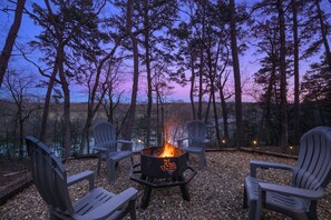 Four empty chairs encircle a lit fire pit in a forested area at dusk, with a pink and purple sky in the background.