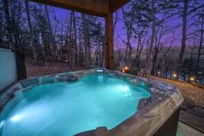 A glowing blue hot tub under a wooden canopy at twilight overlooks a forested area with mountains in the distance and surrounding string lights for ambiance.