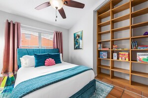 A cozy room with a teal bedspread and coral accents w/ board games on the shelf