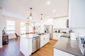 This bright, modern kitchen features white cabinetry, stainless steel appliances, and a light grey island with seating for two. The dining area offers a wooden table for six, with large windows filling the space with natural light.
