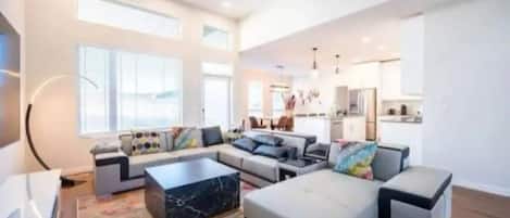This modern living room features a light grey sectional sofa with colorful pillows, a marble coffee table, and a patterned rug. Large windows fill the space with natural light, with a sleek kitchen and dining area in the background.