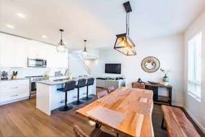 This open-concept kitchen features white cabinets, stainless steel appliances, and an island with seating for three. The dining area has a live-edge wooden table with six chairs and a bench, all set on hardwood floors with industrial pendant lighting