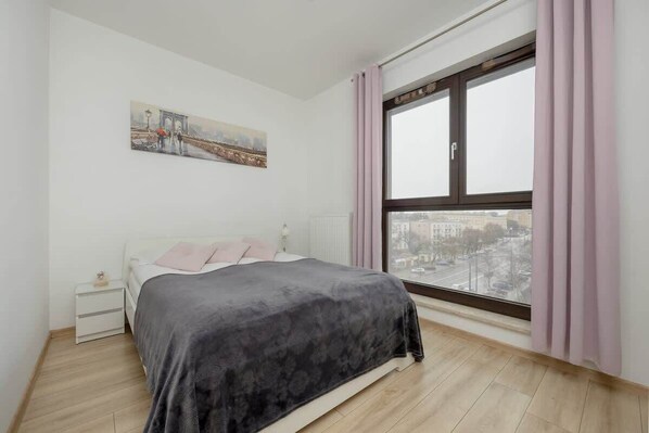 A modern bedroom with a large window, a comfortable bed with dark gray bedding, and soft pink curtains, creating a cozy and elegant atmosphere.

