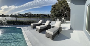 A poolside patio with three cushioned lounge chairs and a table set near a fence overlooking a lake, under a clear blue sky with scattered clouds. Trees and houses are visible in the background.