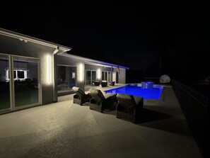 Nighttime view of a modern house's outdoor area with illuminated swimming pool, lounge chairs, and patio furniture.