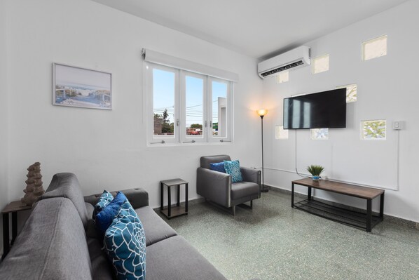 Spacious living room with two large, comfortable sofas, perfect for relaxing. A smart TV is mounted on the wall, and a collection of board games is available on the shelf for entertainment. The room has plenty of natural light from large windows.