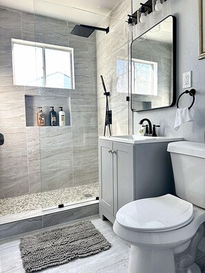 A modern bathroom with a white bathtub, sink, and toilet. The walls are tiled in light gray with a large mirror above the sink.
