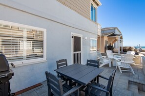 Beachfront patio with a dark gray dining table, Adirondack chairs around a fire pit, and a grill. The ocean is visible in the background, along with neighboring coastal homes. Relaxed outdoor setting perfect for dining and socializing by the beach.