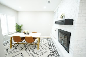 "Gather around this bright, chic dining area that blends classic charm with contemporary style. The white brick fireplace adds a cozy ambiance, perfect for intimate dinners or a coffee chat on a lazy morning. The geometric patterns of the rug underfoot pa