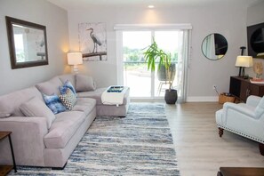 This inviting living room has a gray couch, plush carpet, and a large window framing the balcony view. An indoor plant adds a touch of greenery, complementing the aesthetic decor. Warm light from a lamp sets the perfect ambiance for relaxation.