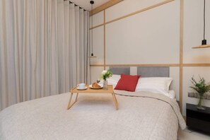 A cozy bedroom with a soft bed, warm lighting, minimalist decor, and a wooden bedside table holding a small plant and lamp.

