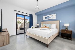 A cozy bedroom with a light bed against a dark blue accent wall, white linens, and light blue pillows. A fish artwork hangs above, and each side has a dark nightstand with lamps that give off a beach vibe!