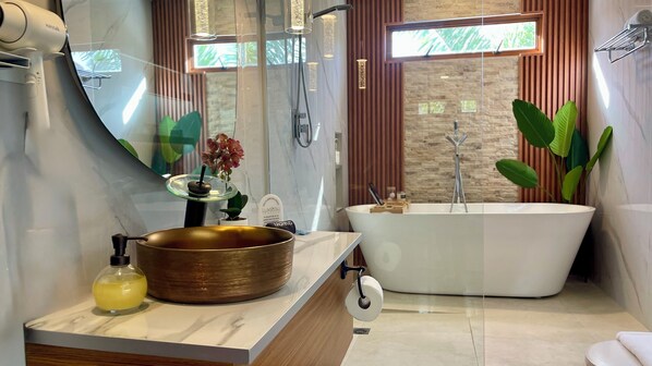 Step into a private spa retreat featuring a soaking tub, rain shower, and gold vessel sink. Surrounded by natural textures and tropical greenery, this serene space blends elegance and comfort with every detail designed for relaxation