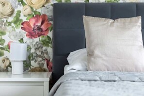 A close-up of a bedside table with a decorative lamp, fresh flowers, and a soft pillow resting against a patterned wall.


