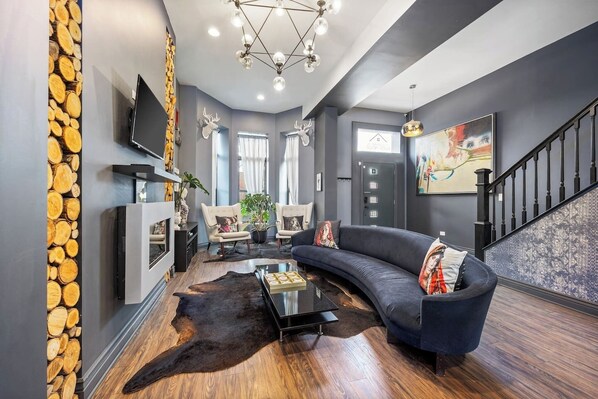 Wide view of the  living room with a fireplace, a plush couch, chairs, and a television, all resting on warm hardwood floors.