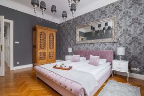 A cozy bedroom with wallpaper, a wooden wardrobe, and a plush bed with pastel bedding.