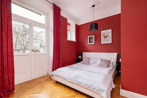 A bright room with bold red walls, a white bed, and a large window. A black pendant lamp adds contrast.